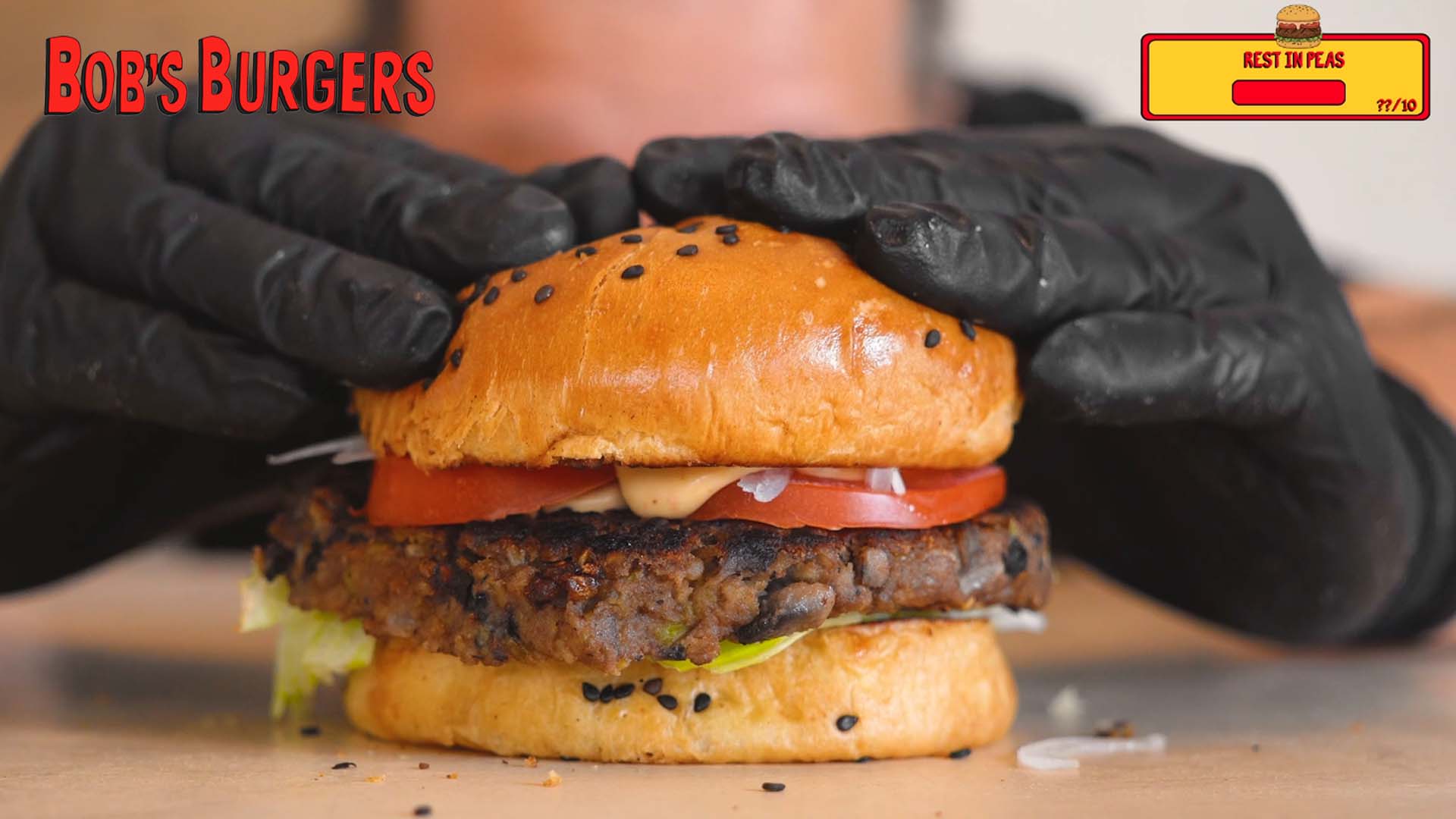 Gloved hands pressing a dark bean patty with tomato into a black sesame bun